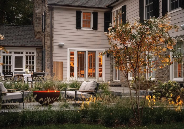 Dusk backyard patio with Corten steel bowl fire pit, modern lounge seating, ornamental grass plantings, multi-stem serviceberry tree, and classic clapboard and fieldstone home exterior