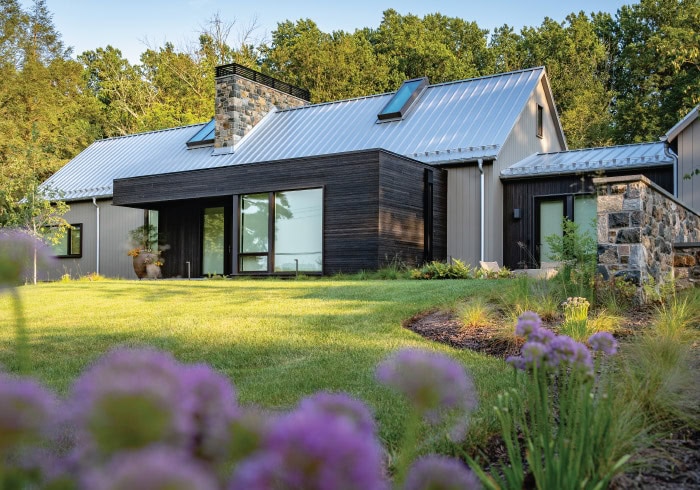 Contemporary farmhouse exterior with standing seam metal roof, charred wood siding, fieldstone chimney, manicured lawn, and allium perennial garden border in foreground