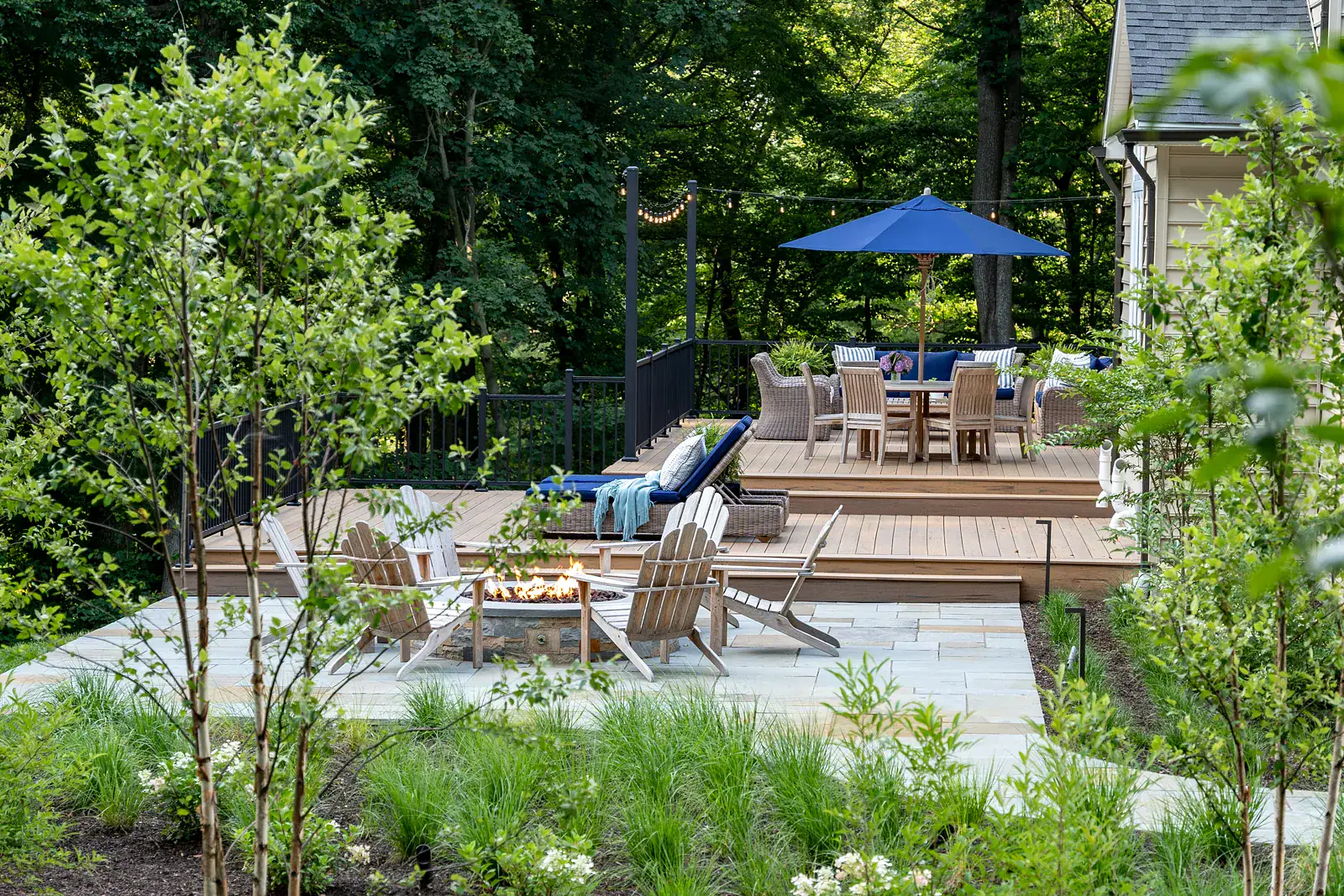 GR-00076 Multi-level residential outdoor living design with bluestone patio, stone fire pit with Adirondack seating, composite deck dining area, multi-stem tree planting, and ornamental grass border