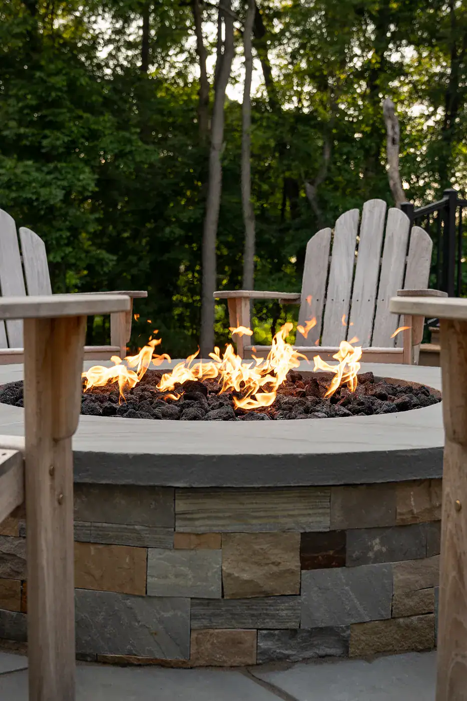 GR-00082 Custom round stone veneer fire pit with lava rock and open flame, surrounded by Adirondack chairs on a residential bluestone patio