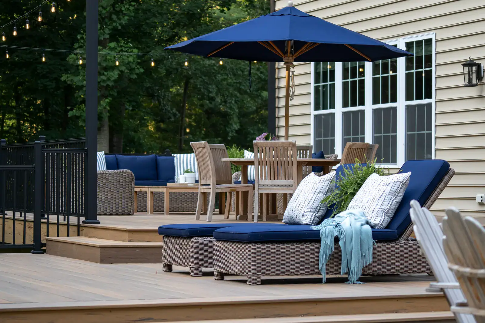 GR-00156 Multi-level composite deck with wicker chaise lounges, teak dining set, wicker lounge seating, patio umbrella, string lights, and black aluminum railing beside a residential home