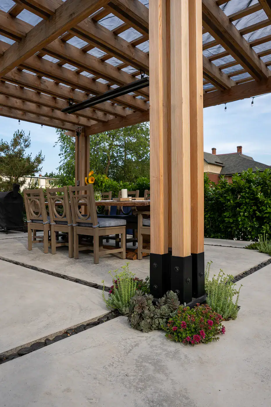 Cedar pergola post detail with steel base hardware, succulent and sedum planting pocket, concrete patio with pebble inlay, and outdoor dining space beneath custom hardscape structure