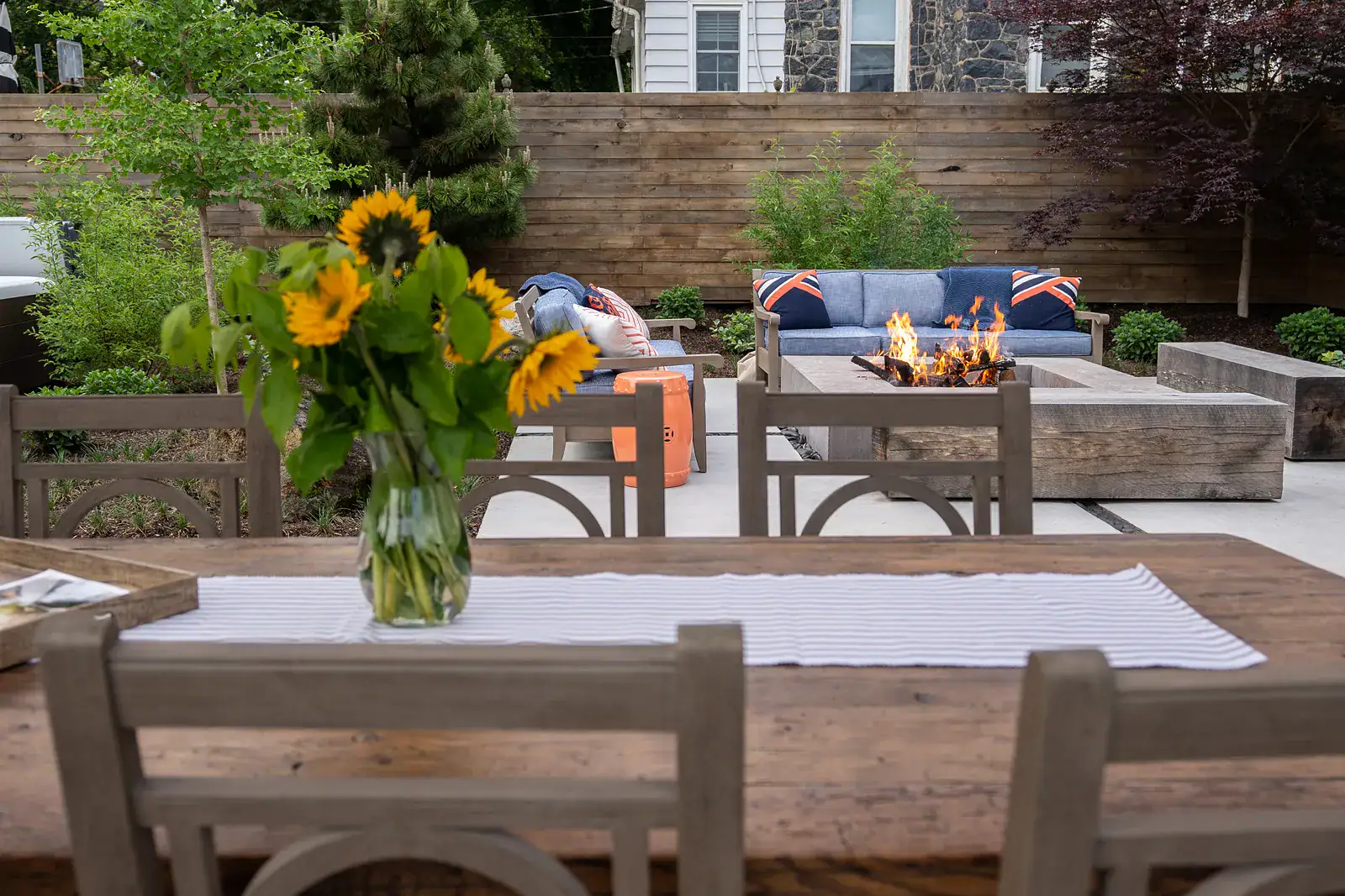 Outdoor dining and lounge patio with concrete fire pit table, outdoor seating, wood privacy fence, and mixed shrub planting border in a custom residential backyard design