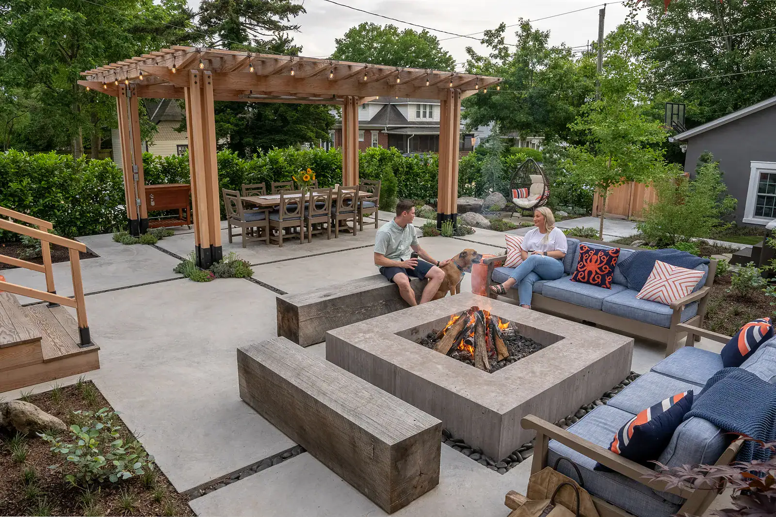 Residential outdoor living space with concrete fire pit table, reclaimed wood bench seating, cedar pergola dining area with string lights, and dense evergreen privacy hedge border