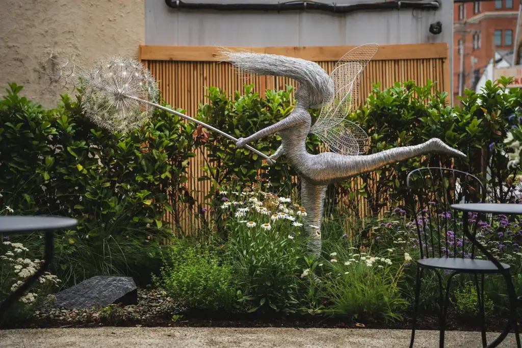 Wire fairy sculpture holding a dandelion seed head in a garden bed of echinacea and verbena with cafe seating alongside