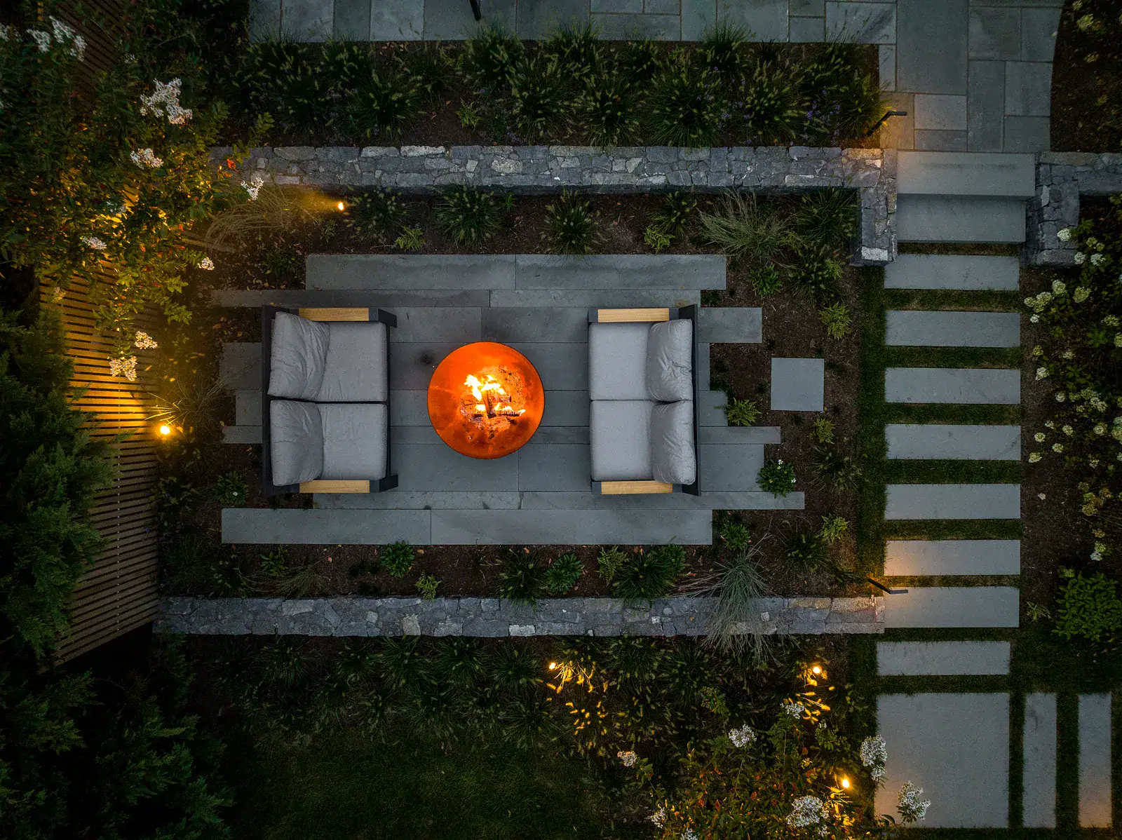 Aerial night view of custom outdoor living patio with lit Corten steel fire bowl, lounge seating, dry-stacked stone retaining walls, stepping stone pathway, and landscape lighting