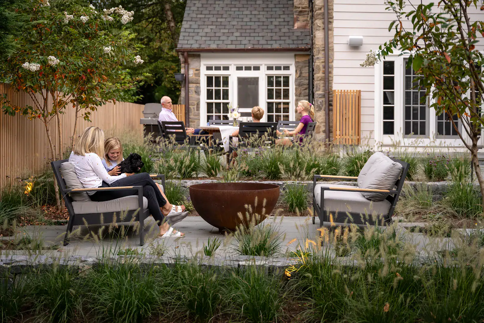 Family gathering on custom residential bluestone patio with Corten steel fire bowl, ornamental grass planting design, and outdoor dining area beside a historic stone and clapboard home
