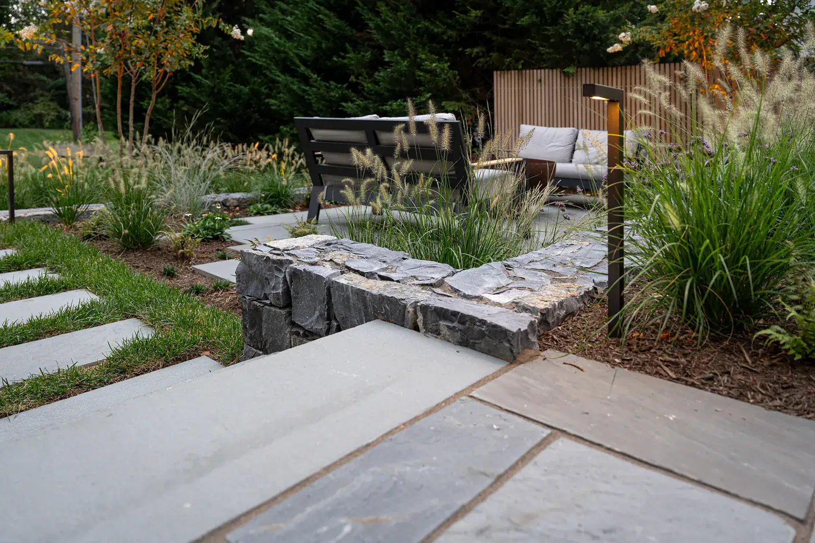 Bluestone walkway with dry-stacked stone retaining wall, ornamental grass planting bed, and custom outdoor living space with cedar privacy screening