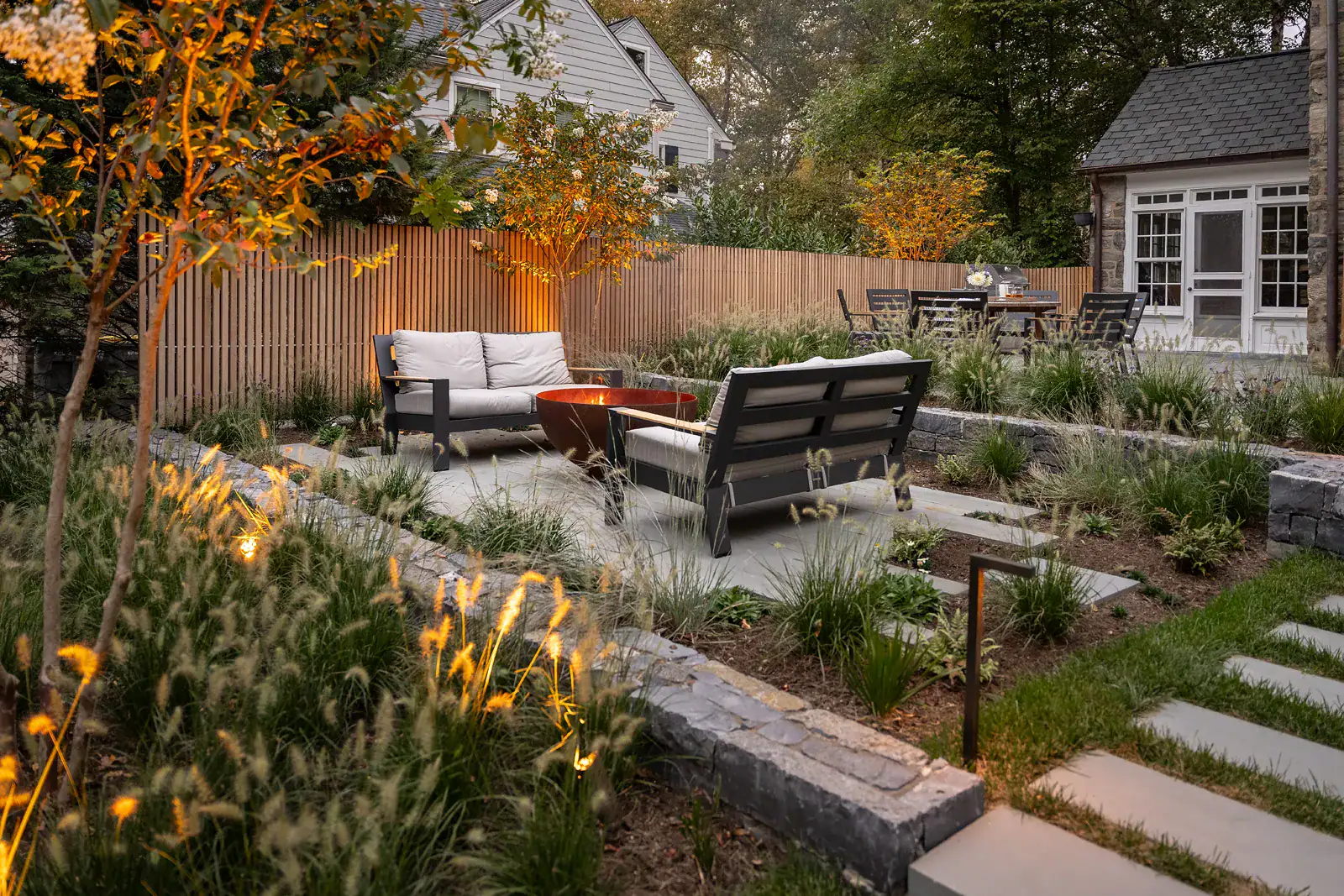 Bluestone patio outdoor living space with Corten steel fire bowl, upholstered lounge seating, ornamental grass planting beds, stone retaining walls, and cedar privacy fence