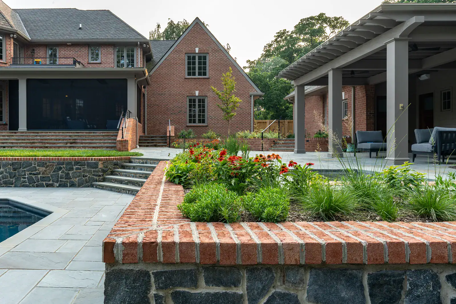 Brick-capped stone retaining wall with echinacea and ornamental grass planting beside a bluestone pool deck and covered outdoor living area in a Wilmington residential landscape