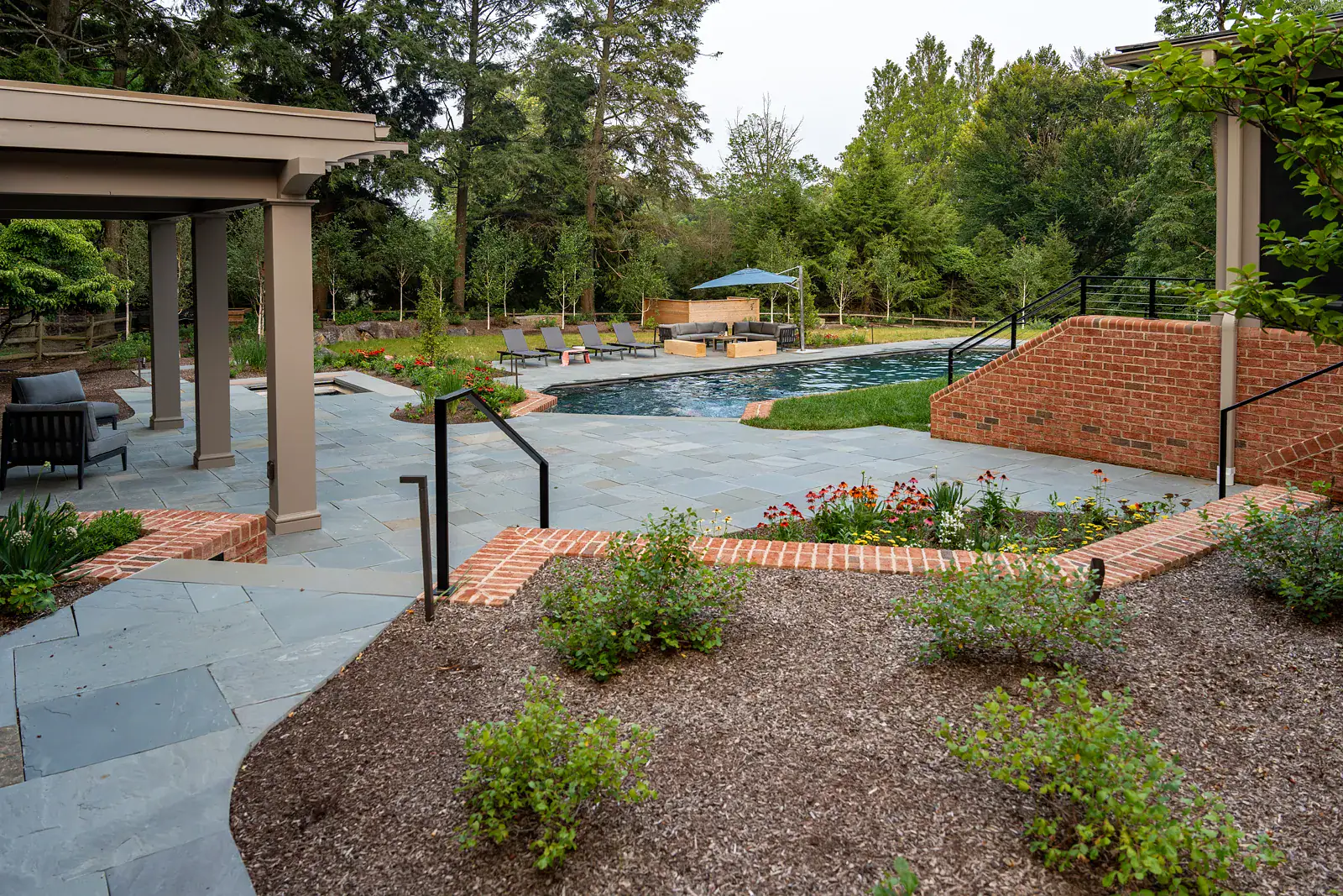 Custom bluestone pool terrace with brick-edged perennial beds, pergola, and outdoor lounge seating surrounded by mature trees