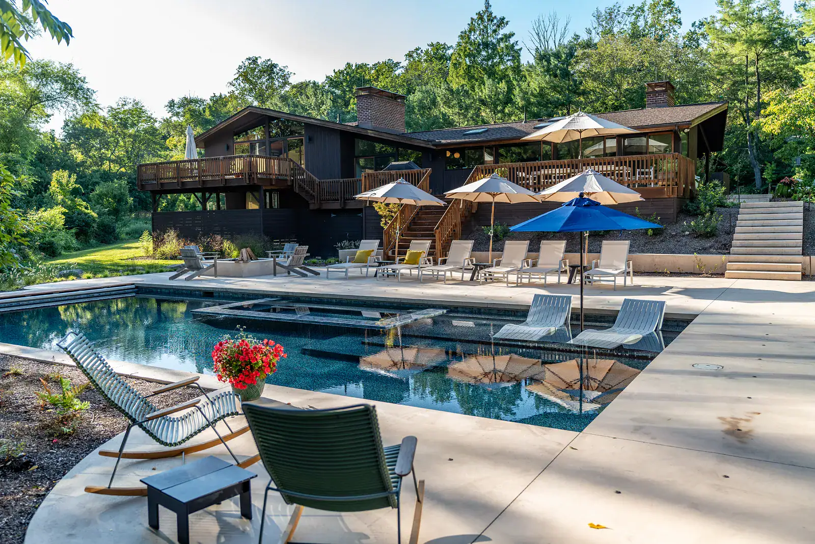 Custom residential pool design with in-pool sun shelf, chaise lounges, market umbrellas, and multi-level wood deck on a mid-century modern home