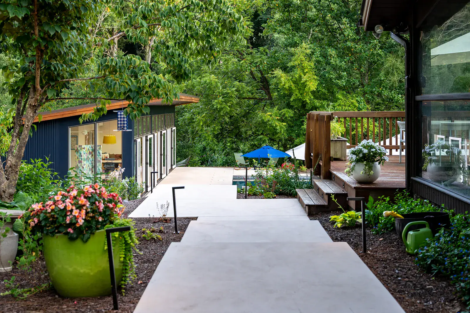 Concrete pathway connecting a modern studio outbuilding to a wood deck with lush shade garden planting, container flowers, and naturalistic landscaping