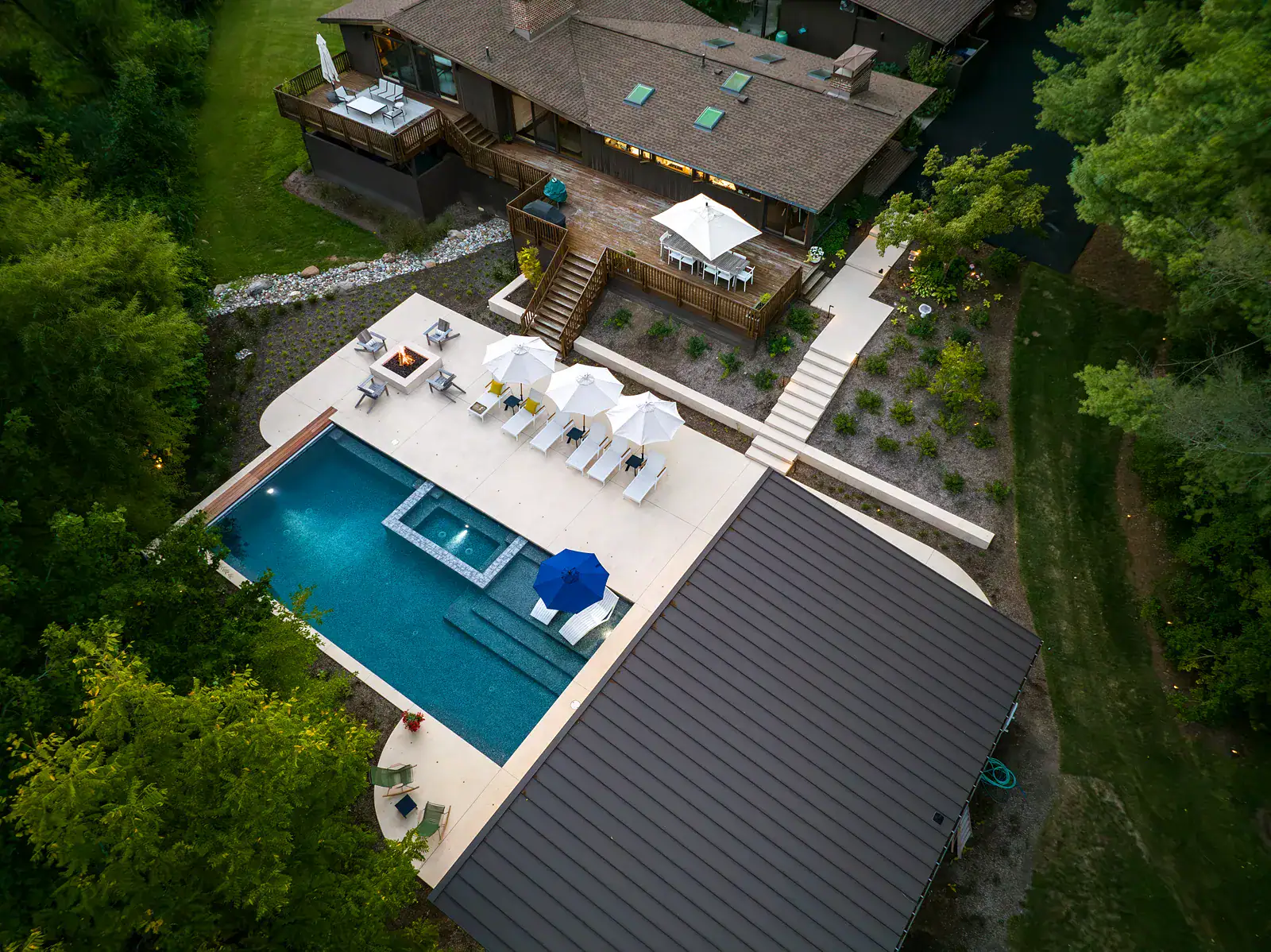Aerial view of a custom residential pool and spa design with concrete deck, fire pit seating area, chaise lounges, wood deck, and naturalistic perennial planting beds