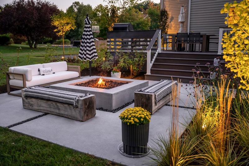 Concrete patio with square concrete fire pit, reclaimed wood bench seating, modern outdoor sofa, ornamental grass planting border, container planting, and composite deck with horizontal rail beyond