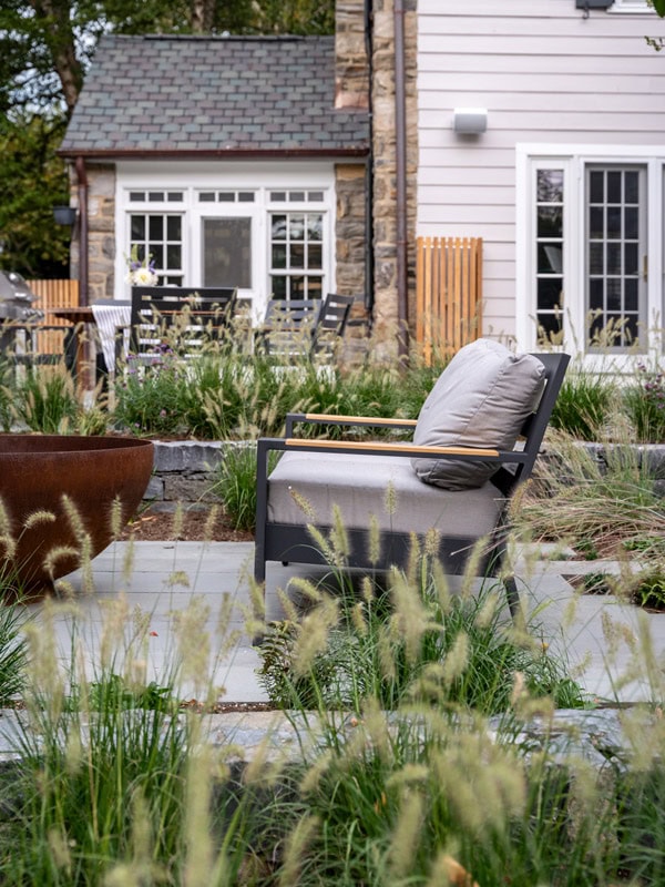 Bluestone patio with modern aluminum and teak lounge chair, Corten steel bowl fire pit, dense ornamental grass planting beds, dining area beyond, and stone and clapboard home exterior