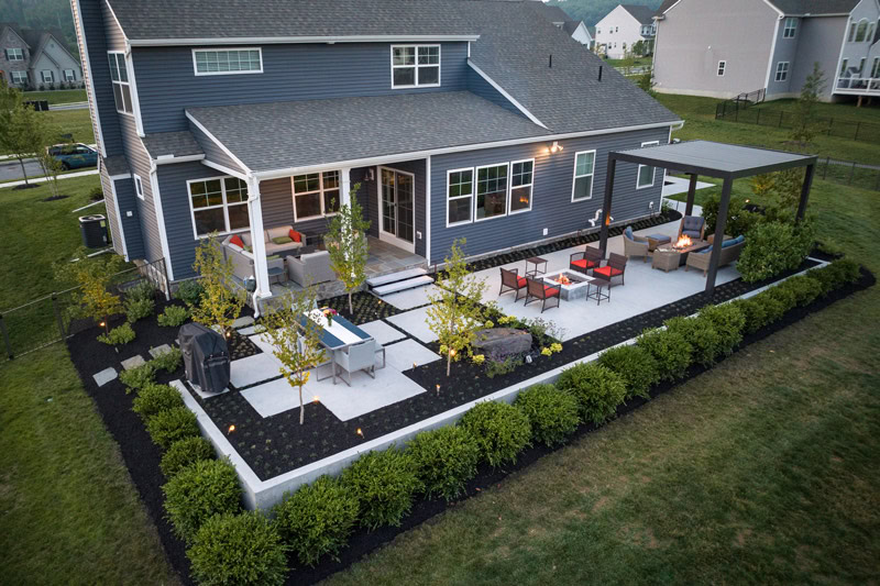 Aerial view of residential backyard landscape design with concrete patio, fire pit lounge area, pergola, outdoor dining, multi-stem trees, shrub border, and landscape lighting