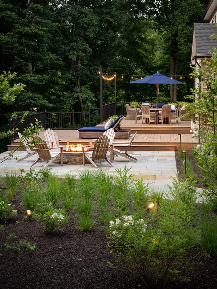 Multi-level cedar deck and bluestone patio with stone fire pit, Adirondack chairs, wicker lounge seating, outdoor dining under navy umbrella, string lights, ornamental grass planting beds, and landscape lighting in a wooded backyard