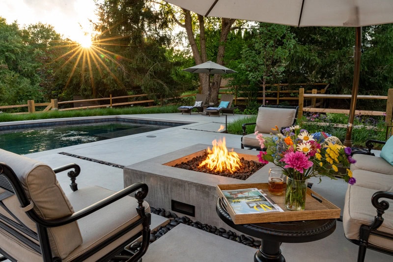 Concrete pool deck with square concrete fire pit, cast iron lounge seating, rectangular pool with spa, chaise lounges, split-rail fence, and wooded backdrop at sunset