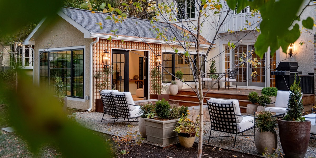Gravel courtyard patio with iron lounge chairs and white cushions, container plantings of boxwood and topiaries, specimen birch tree, and cedar deck with open glass door to a warmly lit interior