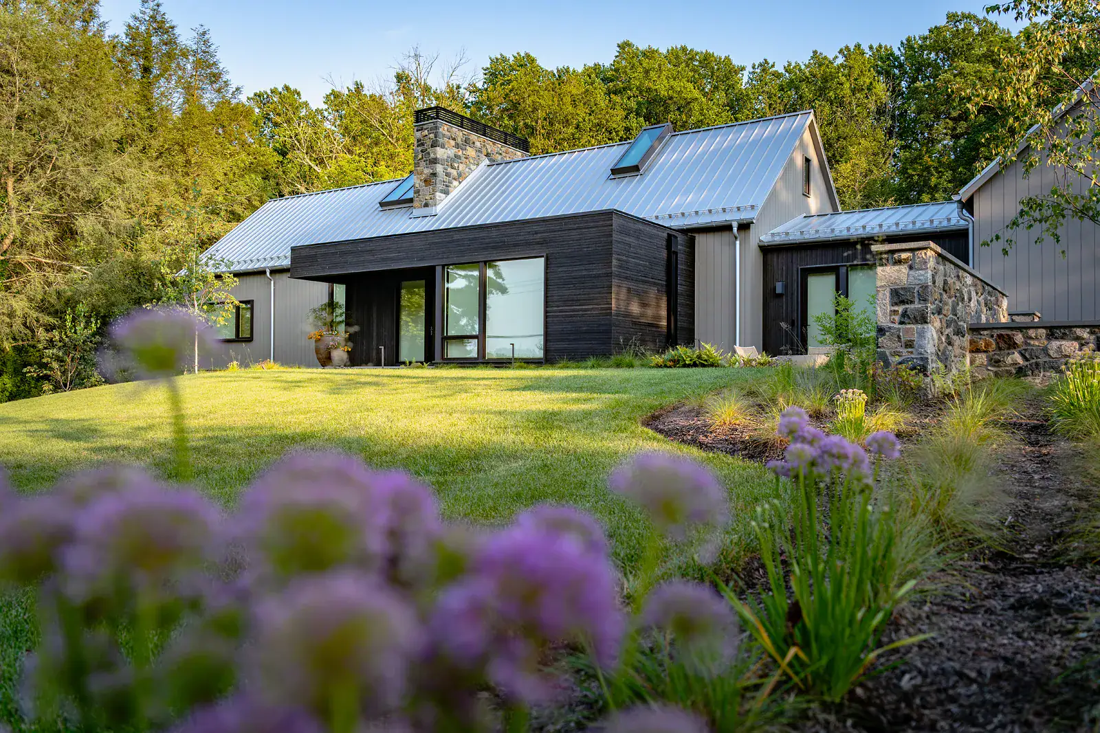 Modern farmhouse rear exterior with metal roof and stone chimney viewed across a lawn with allium blooms and perennial border in the foreground