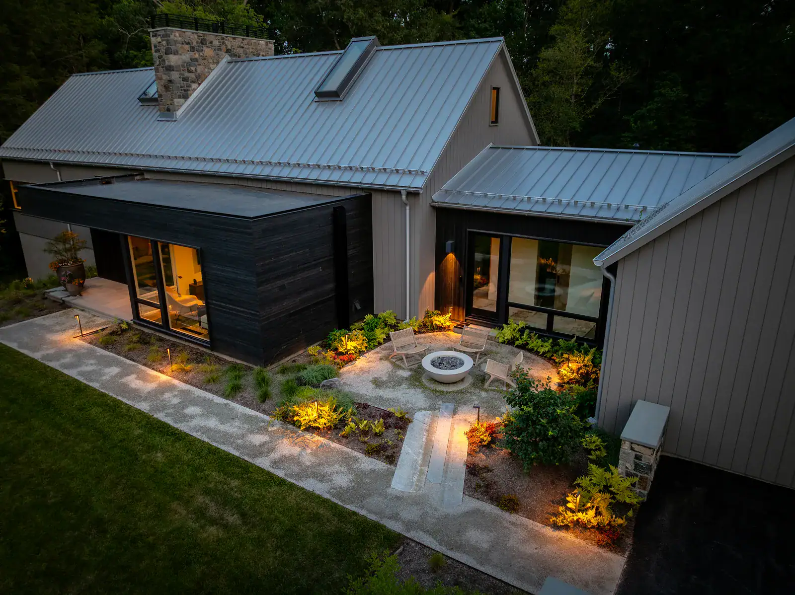 Elevated view of a modern farmhouse courtyard at dusk with circular gravel fire pit seating area, perennial plantings, bluestone steps, and landscape lighting between dark wood and board-and-batten building wings