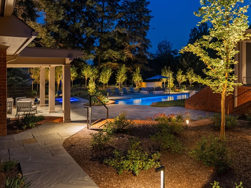 Evening pool terrace with illuminated rectangular pool, covered pergola with dining seating, uplighted ornamental trees, flagstone pathway, perennial planting beds, and bollard lighting at dusk