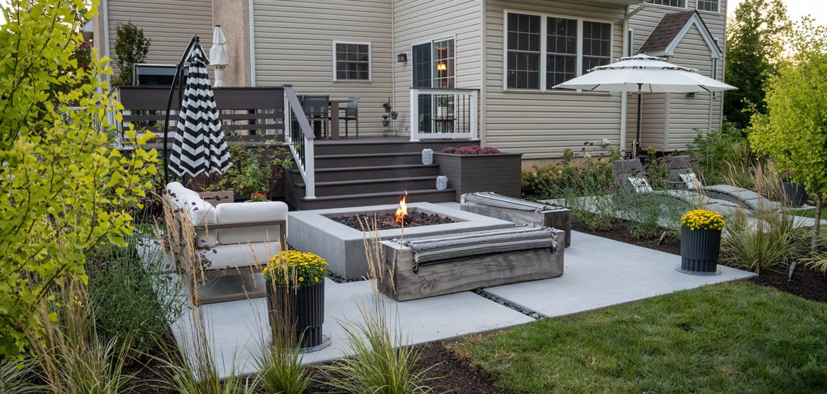 Concrete patio with square concrete fire pit, reclaimed wood bench seating, outdoor lounge chairs with umbrella, container plantings, ornamental grass border, and composite deck with staircase leading to home