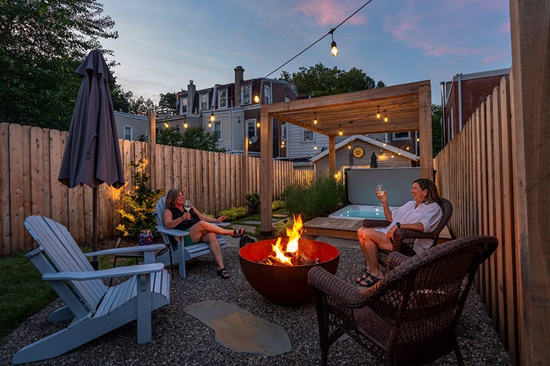 Two women relaxing at dusk around a Corten fire bowl on a gravel patio with Adirondack chairs, string lights, cedar pergola, and hot tub in a small urban backyard design