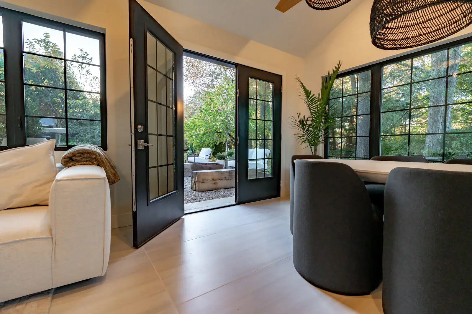 Black-framed French doors opening from an indoor living space to a wooded outdoor patio with a fire feature, part of a Villanova outdoor room designed by GreenRoots