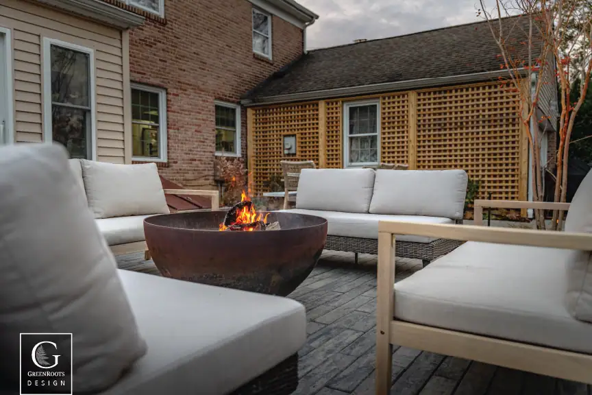 Alt text for this image: Cast iron fire bowl surrounded by cushioned patio seating on a bluestone patio, with a wood lattice privacy screen and Villanova home in the background