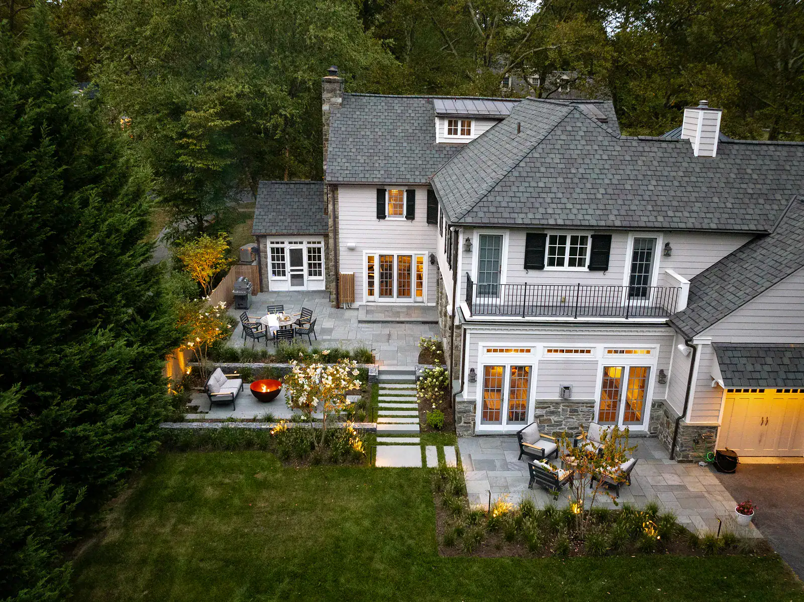 Aerial night view of custom outdoor living patio with lit Corten steel fire bowl, lounge seating, dry-stacked stone retaining walls, stepping stone pathway, and landscape lighting
