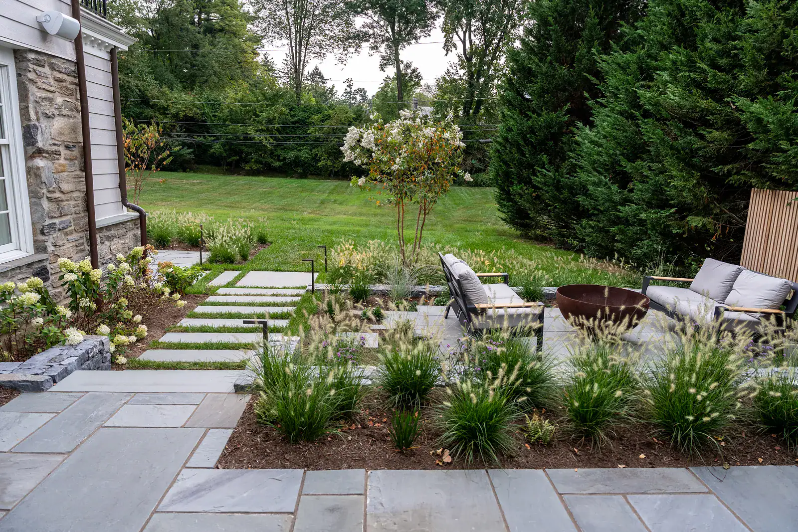 Custom residential landscape design with bluestone patio, stepping stone pathway, ornamental grass planting beds, hydrangea border, Corten steel fire bowl, and open lawn beyond