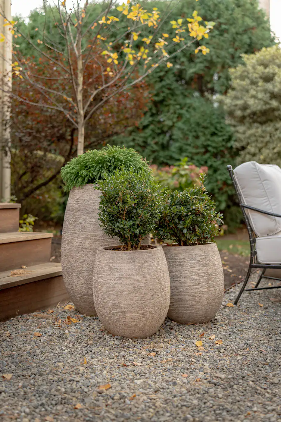 Grouping of textured stone planters with evergreen shrubs on a gravel patio near wood deck steps