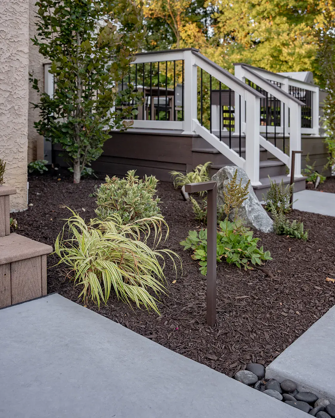 Poured concrete walkway with black river stone joints leading past a variegated hakone grass planting bed, a boulder, a path light, and a columnar tree to a composite deck with white railings and black balusters in a Garnet Valley PA landscape design
