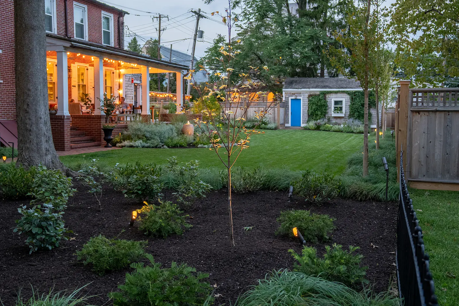 Dusk view across a West Chester PA garden design with a young specimen tree and low-lit evergreen plantings in the foreground, a glowing covered brick porch, a fountain urn, an open lawn, and a blue-doored garden shed beyond
