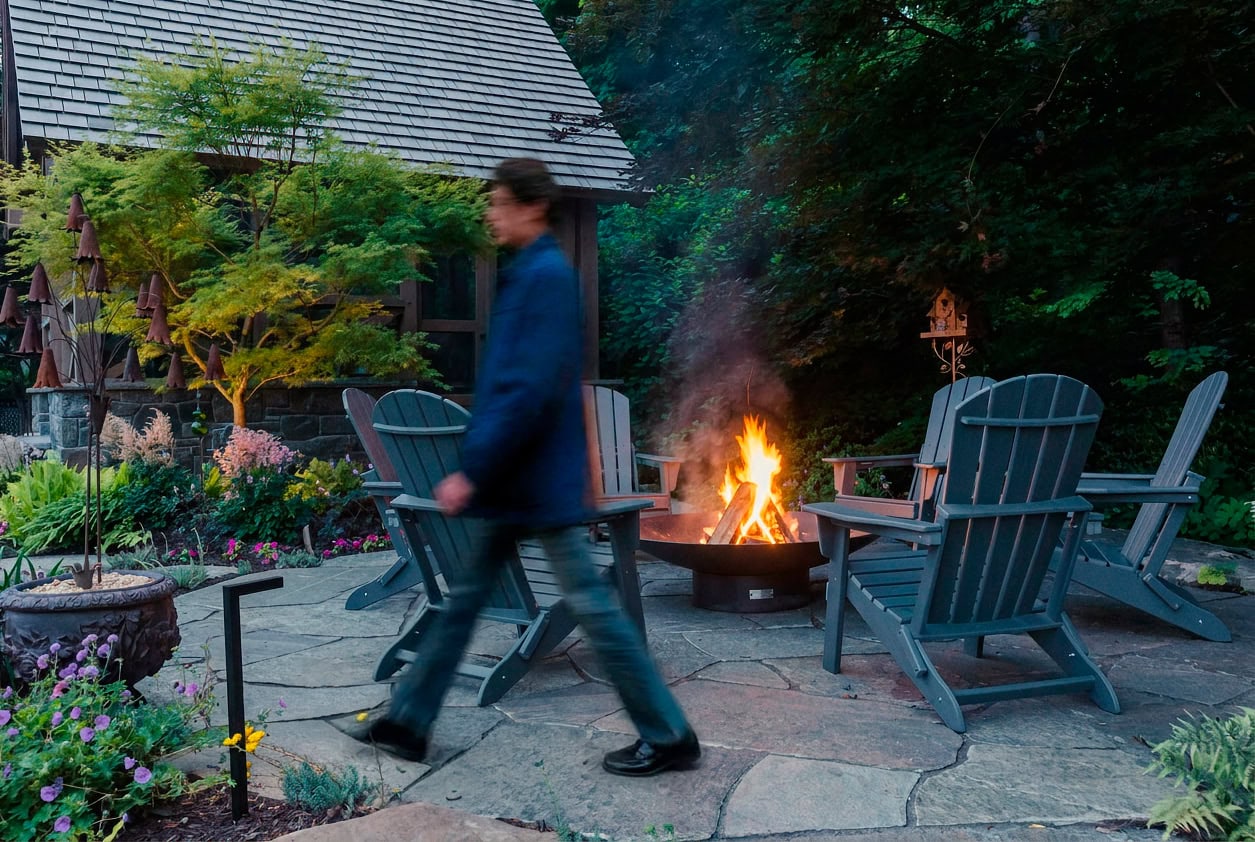 Man walking past a steel fire bowl with a wood fire and gray Adirondack chairs on an irregular flagstone terrace in a Lincoln University PA woodland garden design, with a laceleaf maple, a rusted bell flower sculpture, a planted urn, and the timber-frame screen room beyond