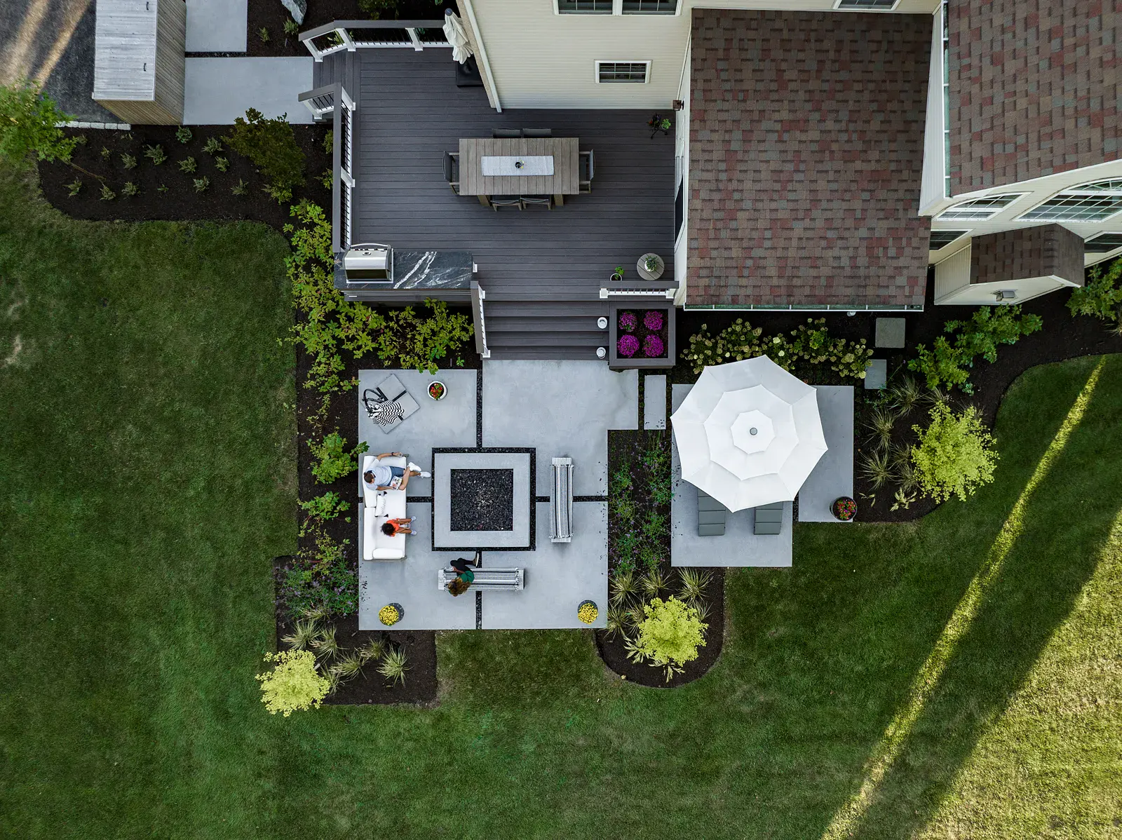 Overhead view of a Garnet Valley PA backyard landscape design with a square concrete fire table, timber benches, and sofa on a poured concrete patio, a chaise lounge pad under an umbrella, a composite deck with outdoor kitchen and dining, and mulched planting beds