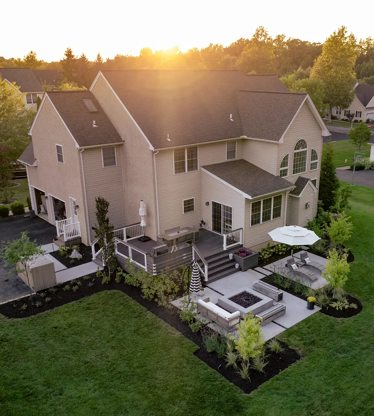 Sunset aerial of a Garnet Valley PA home with a backyard landscape design featuring a composite deck with outdoor kitchen and dining, a concrete fire table patio, a chaise lounge area under an umbrella, small trees, ornamental grasses, and mulched planting beds