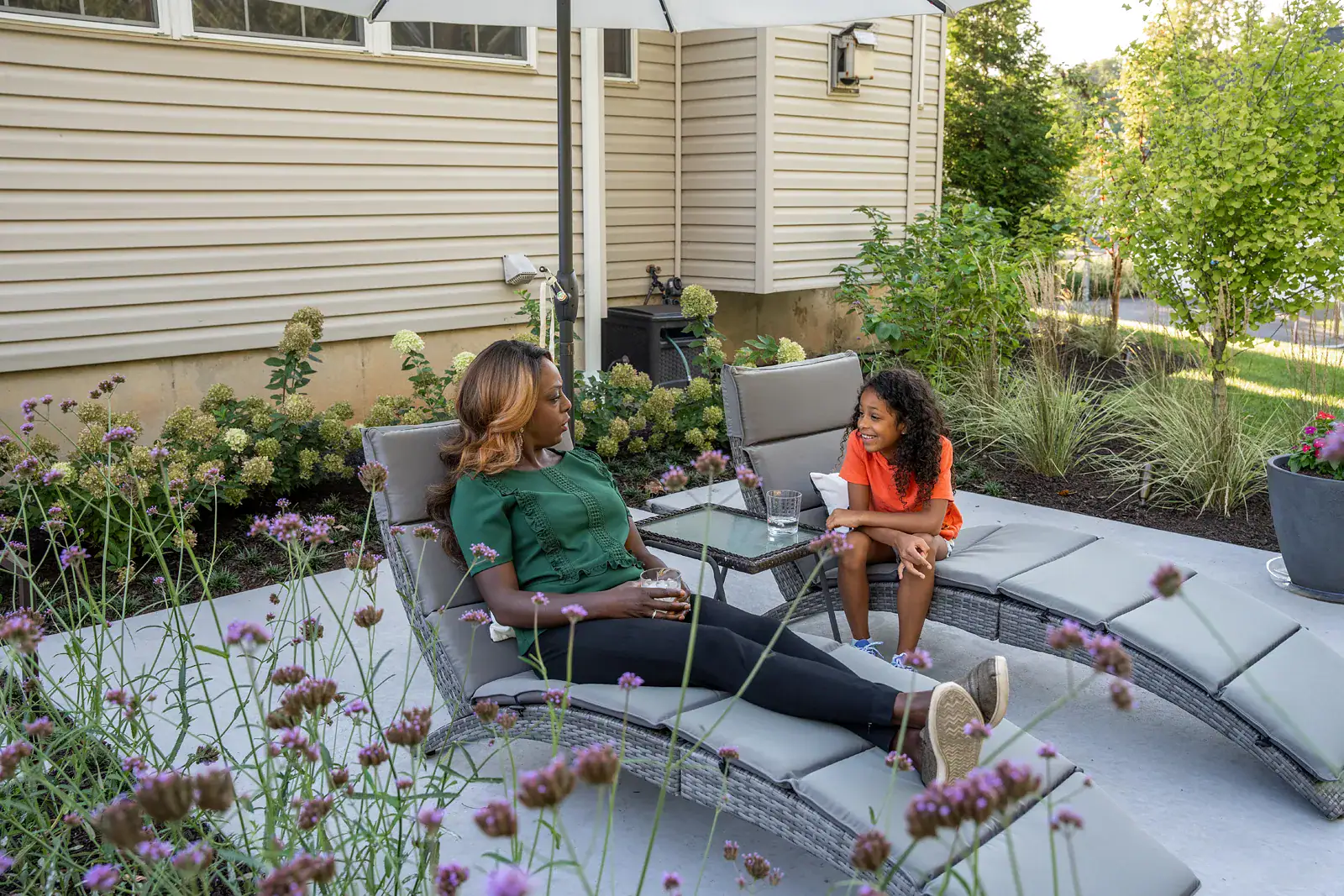 A woman and her daughter talking on gray wicker chaise lounges under an umbrella in a Garnet Valley PA backyard landscape design, with a concrete patio, purple verbena, hydrangeas, ornamental grasses, and a small ginkgo tree