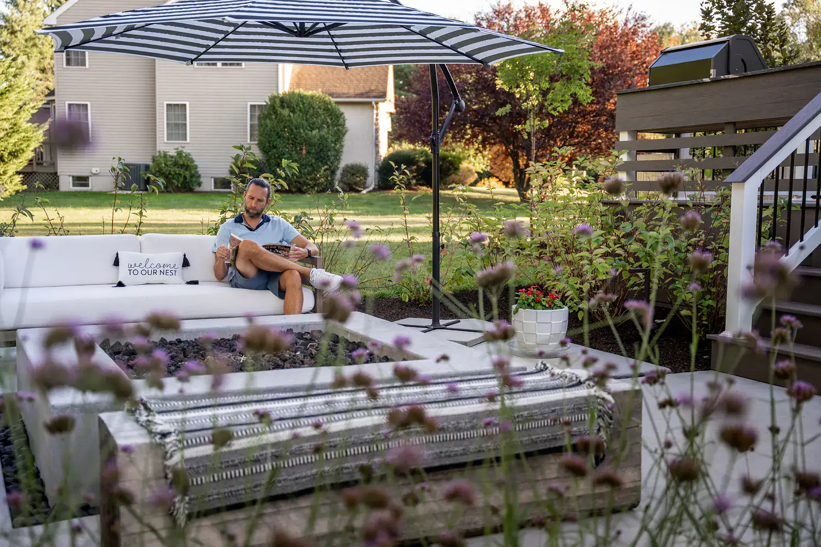 Man reading on a white outdoor sofa beside a concrete fire table in a Garnet Valley PA backyard landscape design, with a striped umbrella, a reclaimed timber bench with a striped throw, purple verbena in the foreground, and open lawn beyond