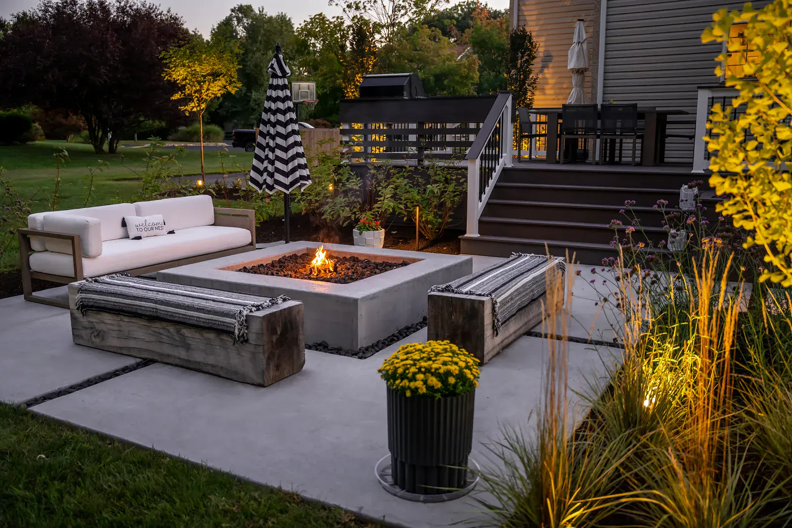 Glowing concrete fire table with two reclaimed timber benches and a white outdoor sofa on a poured concrete patio in a Garnet Valley PA backyard landscape design, with a potted yellow mum, uplit ornamental grasses, a striped umbrella, and a composite deck beyond