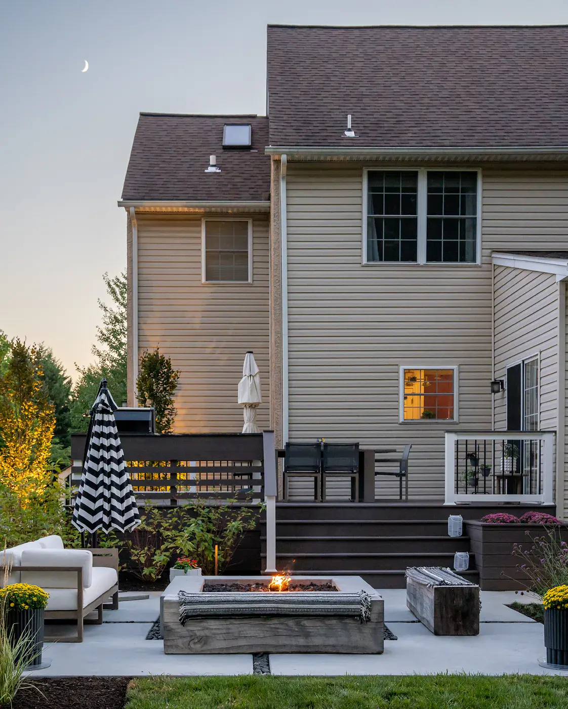 Dusk view of a Garnet Valley PA backyard landscape design with a lit concrete fire table, reclaimed timber bench, and outdoor sofa on a poured concrete patio, a striped umbrella, potted yellow mums, and a composite deck with steps and outdoor dining above