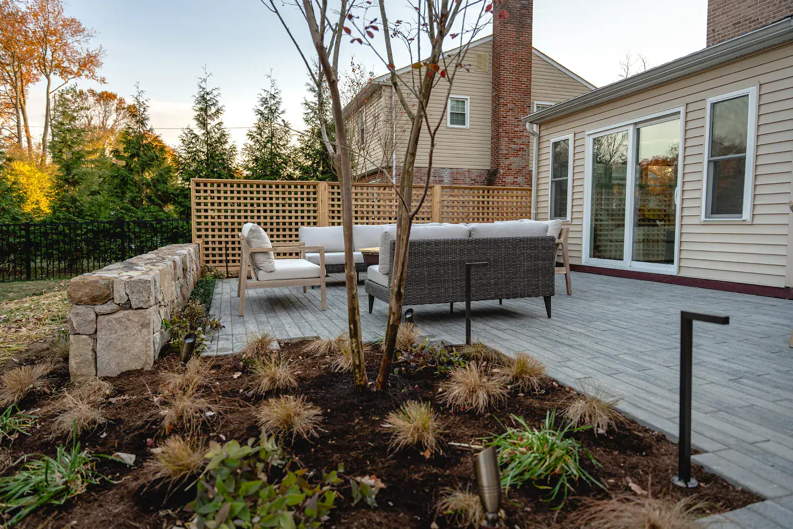Outdoor lounge seating on a gray paver patio in a Wilmington DE landscape design, framed by a dry-stacked fieldstone wall, cedar lattice privacy screening, evergreen backdrop, multi-stem trees, and ornamental grass beds with landscape lighting