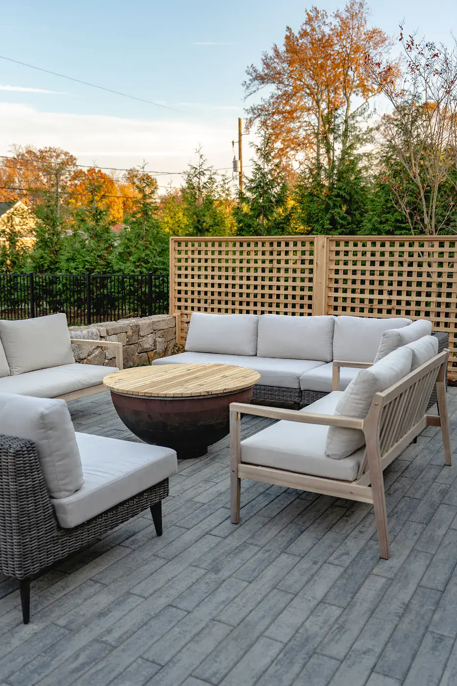 Fireside lounge seating around a steel fire bowl with a wood slat lid on a gray paver patio in a Wilmington DE landscape design, with cedar lattice screening, a fieldstone wall, evergreens, and autumn foliage beyond
