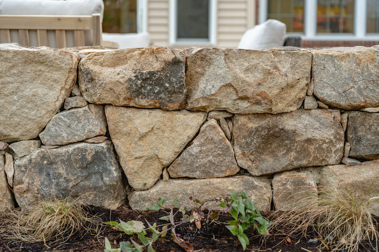 Close detail of a dry-stacked fieldstone seating wall in warm tan and gray tones in a Wilmington DE landscape design, with ornamental grasses and new perennial plantings at its base and patio seating beyond