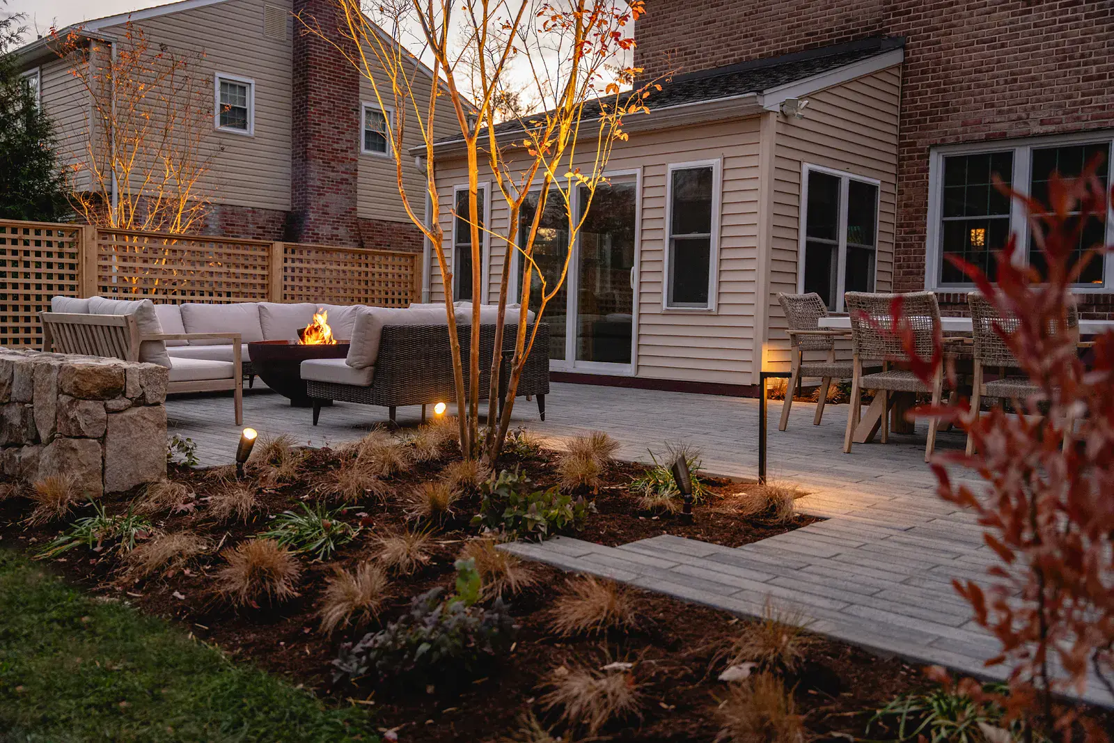 Dusk view of a Wilmington DE landscape design with a lit fire bowl and lounge seating, an outdoor dining area on a gray paver patio, uplit multi-stem crape myrtle, ornamental grass beds, a fieldstone wall, and cedar lattice screening