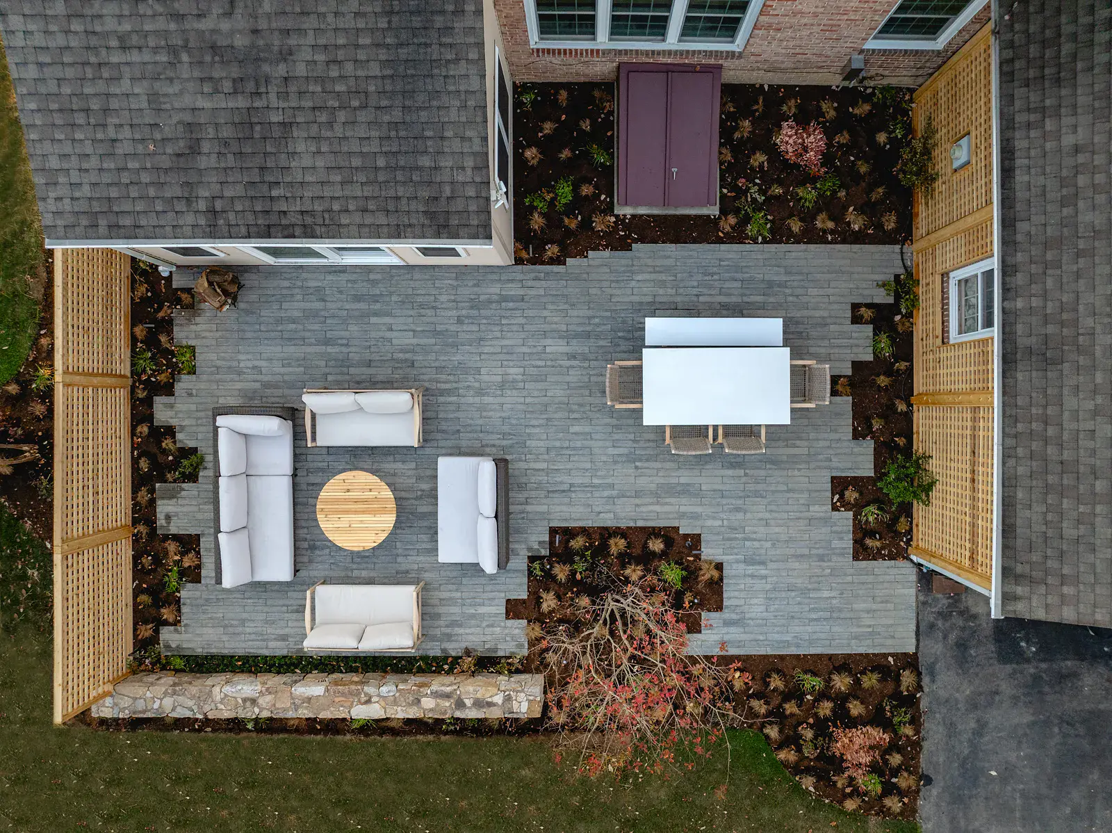 Overhead view of a Wilmington DE landscape design with a gray paver patio, fireside lounge seating around a round fire bowl, an outdoor dining table, cedar lattice screening, a fieldstone wall, and mulched ornamental grass planting beds