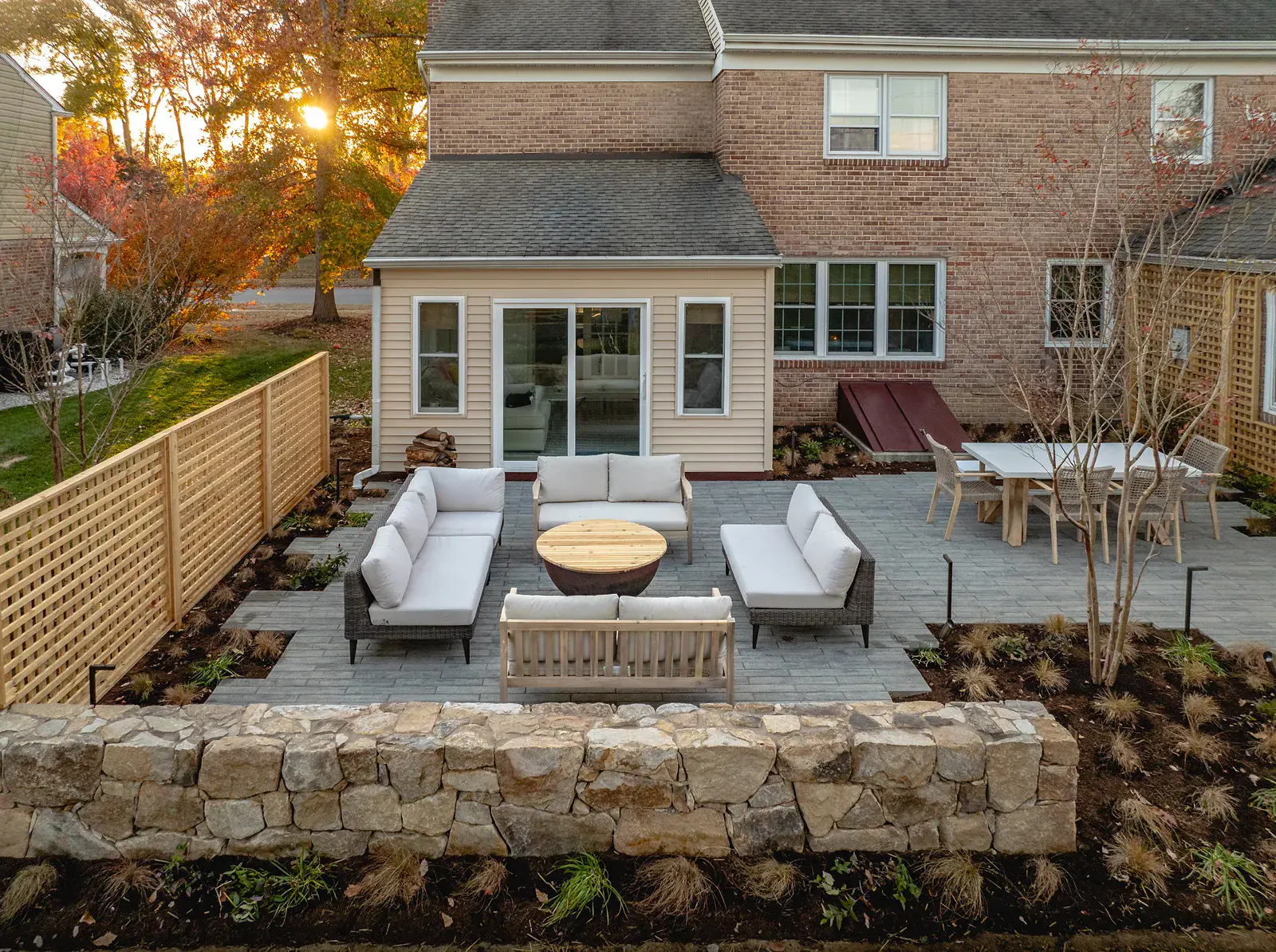 Fireside patio at sunset in a Wilmington DE landscape design, with a steel fire bowl and lounge seating on a gray paver patio, a dry-stacked fieldstone wall, cedar lattice screening, an outdoor dining area, and ornamental grass plantings with autumn foliage
