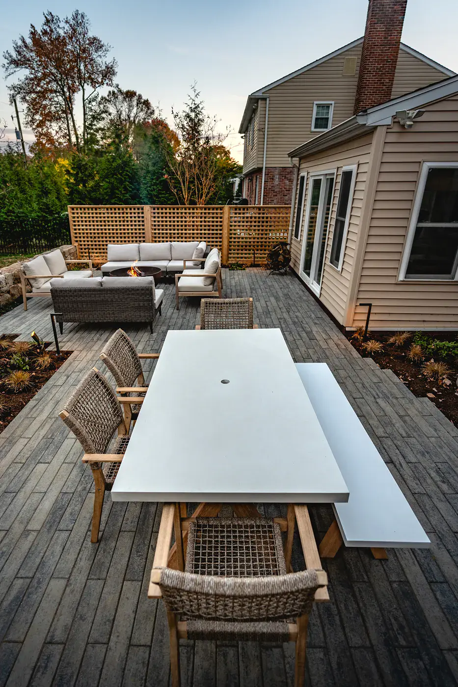 Outdoor dining area with a white concrete table, bench, and woven teak chairs on a gray paver patio in a Wilmington DE landscape design, with a lit fire bowl and lounge seating, cedar lattice screening, and evergreens at dusk